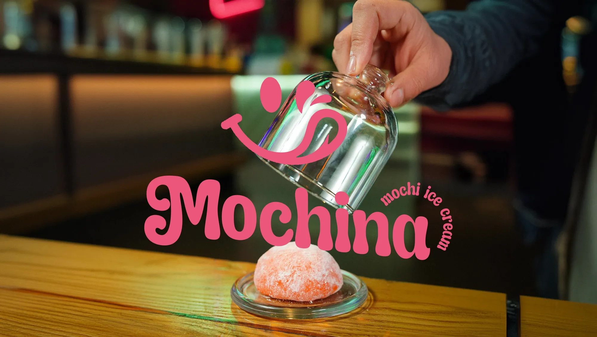 A hand lifting a glass cloche off a pink mochi, with the Mochina logo